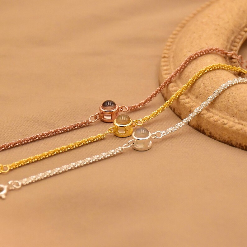May include: Three delicate chain bracelets in rose gold, yellow gold, and silver tones. Each bracelet features a single small, round, bezel-set charm with a smooth, translucent center stone, connected to a fine, textured link chain design.