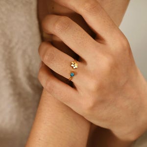 May include: A gold-toned open ring featuring a small paw print charm on one end and a tiny, bright blue heart-shaped gemstone on the other. The band is thin and metallic, designed to wrap partially around the finger with an open gap between the ends.