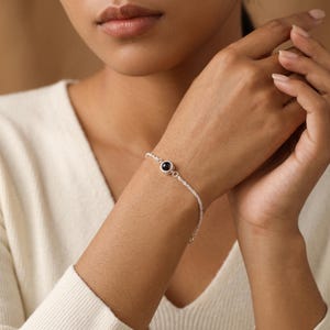 May include: A delicate silver chain bracelet featuring a single, small, round, black stone set in a polished silver bezel. The bracelet has a fine, twisted link design that wraps around the wrist, secured with a simple, functional metal clasp.