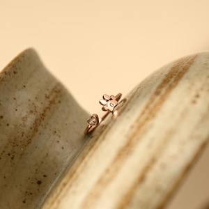 May include: A rose gold open ring featuring a small paw print charm with the letter G engraved on the pad and a tiny heart-shaped charm on the opposite end of the band. The ring has a polished metallic finish and a delicate, adjustable design.