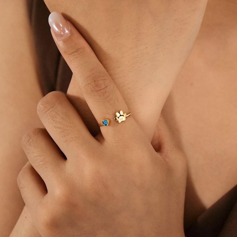 May include: A gold-toned open ring featuring a small blue heart-shaped stone on one end and a gold paw print charm with the letter W engraved on the other end. The ring is designed with a thin, adjustable band that wraps around the finger.