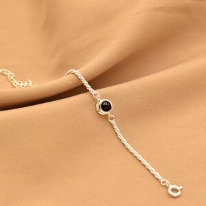 May include: A delicate silver-toned chain bracelet featuring a single, small, round, black stone set in a polished silver-toned bezel. The bracelet includes a thin, textured chain and a small circular clasp at the end for secure fastening.