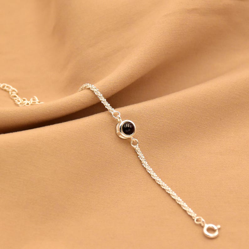 May include: A delicate silver-toned chain bracelet featuring a single, small, round, black stone set in a polished silver-toned bezel. The bracelet includes a thin, textured chain and a small circular clasp at the end for secure fastening.