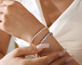 May include: Two tennis bracelets, one gold and one silver, are worn on a wrist. The bracelets are adorned with numerous small, sparkling stones. The background is a white garment.