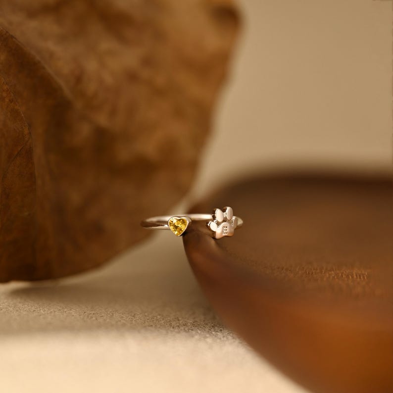 May include: A silver-toned ring featuring a small yellow heart-shaped gemstone on one end and a silver paw print design on the other end. The ring has an open band design, showcasing the contrasting yellow stone and the metallic paw print detail.
