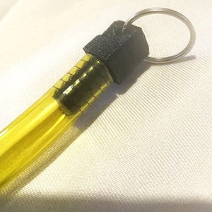 Puede incluir: Un tubo de vidrio amarillo con una tapa negra y un llavero plateado. El tubo está lleno de un líquido transparente.