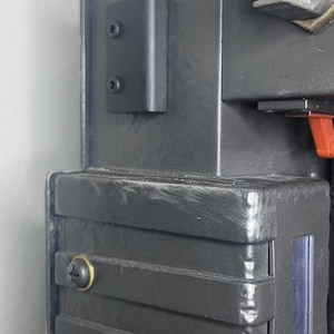 May include: A close-up of a black, rectangular object with a boxy design. The object has a series of horizontal ridges and is secured with screws. A small, black rectangular piece is attached to the side with two screws. The object appears to be a prop or a piece of equipment.