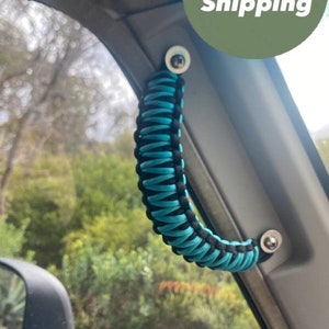 Puede incluir: Un asa de paracord turquesa y negro instalada en un coche. El asa está unida al techo del coche cerca de la ventana.