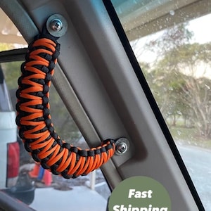 Puede incluir: Mango de cuerda trenzada negro y naranja instalado en un coche. El mango está fijado al interior del coche con dos tornillos plateados. El texto "Envío rápido" es visible en la imagen.