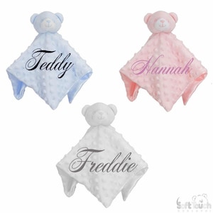 Può includere: Tre coperte per bambini personalizzate con un orsacchiotto attaccato. Le coperte sono blu, rosa e bianche con i nomi "Teddy", "Hannah" e "Freddie" ricamati.