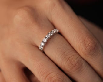 May include: A silver ring with a row of clear, round-cut gemstones. The ring is on a finger, with the skin tone appearing light. The gemstones are evenly spaced and sparkle against the silver band.