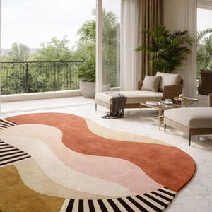 May include: A large, oval area rug with a wavy design in shades of rust, pink, beige, and gold. The rug has a black and white striped border. The rug is placed in a bright, airy room with a balcony.