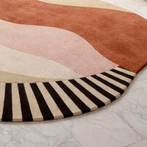 May include: A round rug with a wave design in shades of rust, pink, beige, and cream. The rug's edge features a border of alternating black and cream stripes. The rug is placed on a marble tile floor.