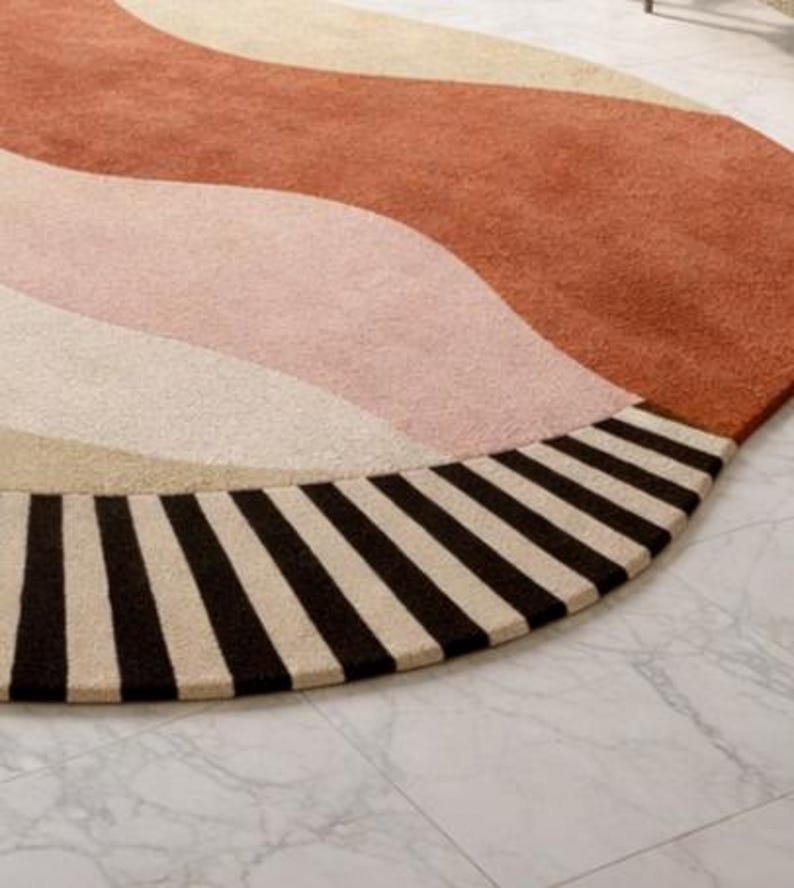 May include: A round rug with a wave design in shades of rust, pink, beige, and cream. The rug's edge features a border of alternating black and cream stripes. The rug is placed on a marble tile floor.