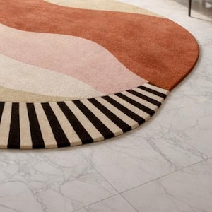 May include: A round rug with a retro design. The rug features a wavy pattern in shades of rust, pink, and cream, with a border of alternating black and cream stripes. The rug is placed on a white marble floor.