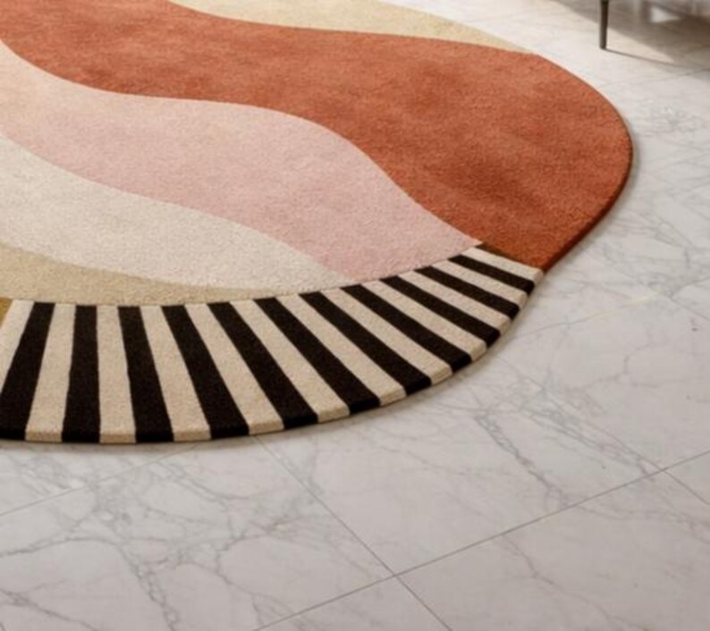 May include: A round rug with a retro design. The rug features a wavy pattern in shades of rust, pink, and cream, with a border of alternating black and cream stripes. The rug is placed on a white marble floor.