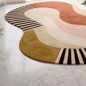 May include: A large, abstract area rug with a wavy design. The rug features a combination of colors, including brown, tan, pink, and gold. The edge of the rug has a striped pattern in black and tan.