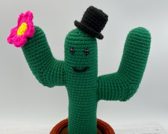 May include: A green crocheted cactus wearing a black top hat and holding a pink flower. The cactus is in a brown and orange pot.