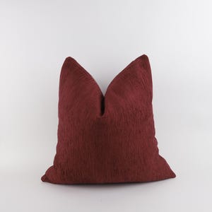 Op de afbeelding: Een bordeauxrood sierkussen met een textuur, geribbelde stof. Het kussen is vierkant en heeft een puntige bovenkant. De achtergrond is effen wit.