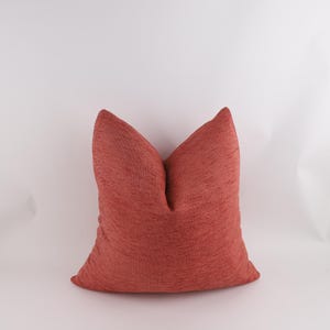 Op de afbeelding: Een vierkant, getextureerd kussen in een warme, roodachtig-oranje tint. Het kussen heeft een zachte, uitnodigende uitstraling, met een subtiel geweven patroon. Het kussen is een decoratief item, geschikt voor een woon- of slaapkamer.