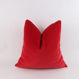 Könnte beinhalten: Ein quadratisches rotes Samtkissen. Das Kissen hat ein weiches, strukturiertes Aussehen und eine klassische Form, ideal, um jedem Wohnraum einen Hauch von Luxus zu verleihen. Die leuchtend rote Farbe macht es zu einem auffälligen dekorativen Akzent.