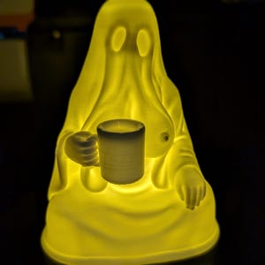 May include: A translucent yellow, glowing ghost-shaped lamp featuring a figure with two oval eyes, holding a small white cylindrical mug in its right hand. The figure has a smooth, draped form and emits a warm, bright light from within.