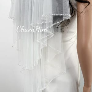 Puede incluir: Un velo de novia blanco con un borde festoneado, adornado con pequeñas cuentas brillantes. El velo es en capas y transparente, con el texto "ChuenHim" impreso en la tela. El velo está diseñado para una boda.