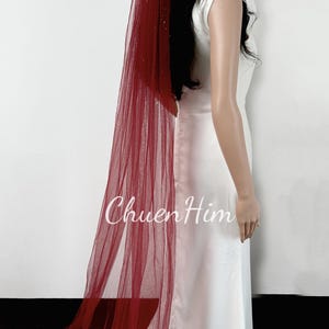 May include: A red tulle wedding veil with a sparkly finish. The veil is long and flowing, and it is worn by a person in a white dress.