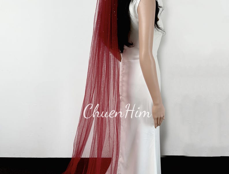 May include: A red tulle wedding veil with a sparkly finish. The veil is long and flowing, and it is worn by a person in a white dress.