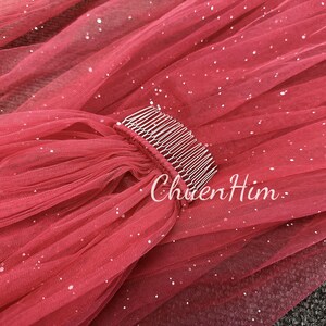 May include: A red tulle veil with silver glitter and a silver comb attached. The veil is gathered at the top and has a flowing design.