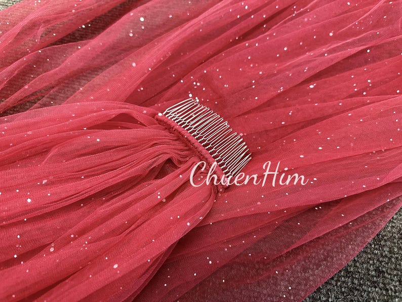 May include: A red tulle veil with silver glitter and a silver comb attached. The veil is gathered at the top and has a flowing design.