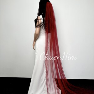 May include: A red tulle wedding veil with a sparkly finish. The veil is long and flows behind the model.