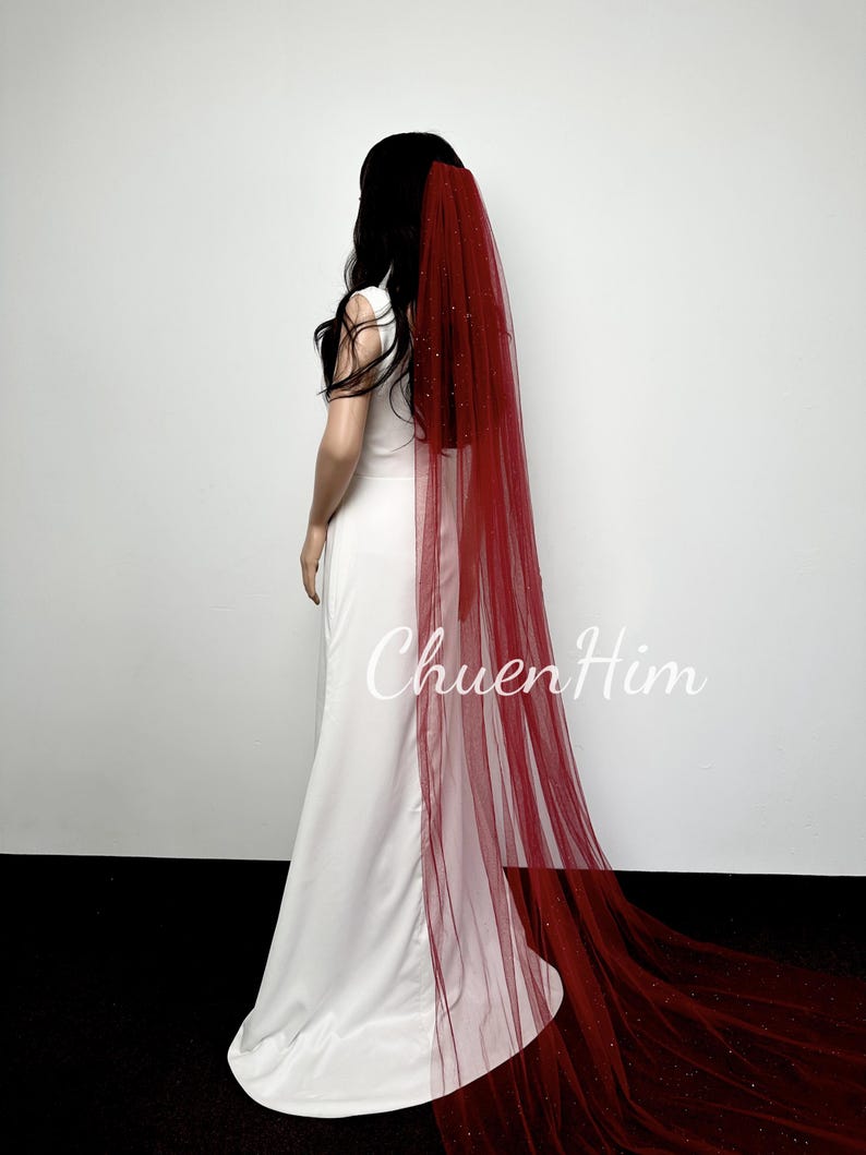 May include: A red tulle wedding veil with a sparkly finish. The veil is long and flows behind the model.