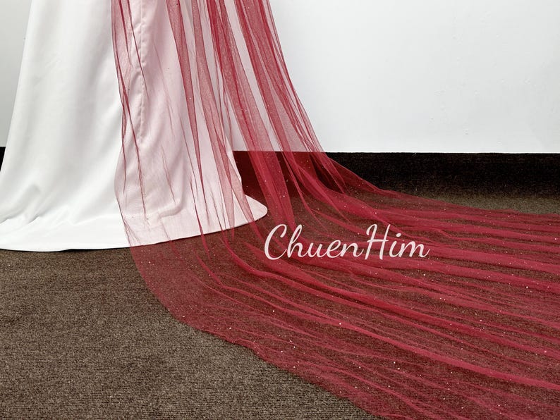 May include: A long, burgundy tulle veil with a sparkly finish. The veil is draped over a white dress.
