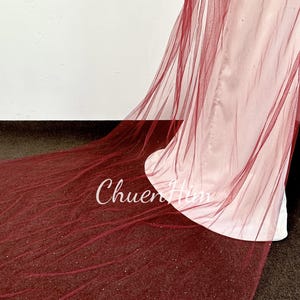 May include: A long, burgundy tulle veil with a sparkly finish. The veil is draped over a white surface.