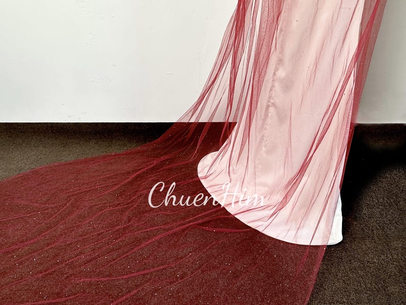 May include: A long, burgundy tulle veil with a sparkly finish. The veil is draped over a white surface.