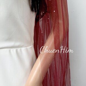 May include: A burgundy tulle shawl with silver glitter accents. The shawl is draped over a person's arm and shoulder.