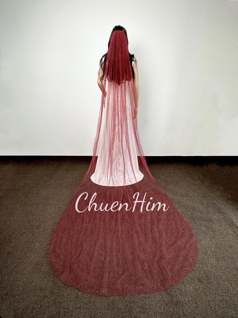 May include: A burgundy bridal veil with a sparkling finish. The veil is made of tulle and has a single layer. The veil is shown on a model wearing a white dress.