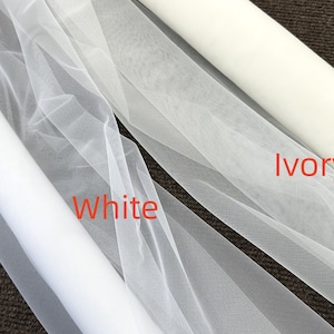 Op de afbeelding: Close-up van twee rollen stof: witte en ivoorkleurige tule. De witte tule is gemarkeerd met "White" in rode tekst en de ivoorkleurige tule is gemarkeerd met "Ivory". De stof is licht en transparant, geschikt voor bruids- of knutselprojecten.
