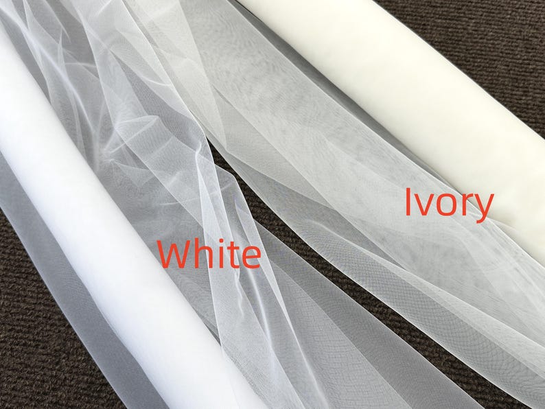 Op de afbeelding: Close-up van twee rollen stof: witte en ivoorkleurige tule. De witte tule is gemarkeerd met "White" in rode tekst en de ivoorkleurige tule is gemarkeerd met "Ivory". De stof is licht en transparant, geschikt voor bruids- of knutselprojecten.