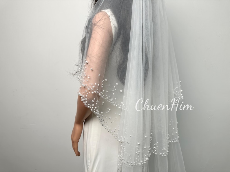 Op de afbeelding: Een witte bruidssluier met twee lagen, versierd met delicate parels langs de randen en op de mouw. De transparante sluier heeft de witte tekst "ChuenHim". Een klassiek bruiloftsaccessoire.