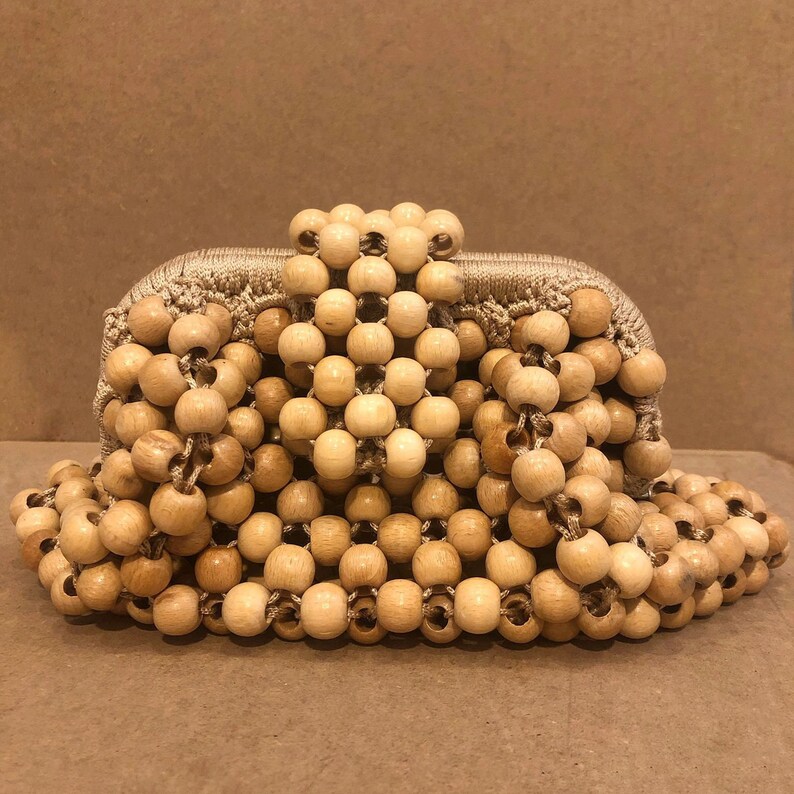 May include: A beige woven clutch bag made entirely of wooden beads. The beads are all the same size and shape, and they are arranged in a pattern that creates a textured surface.