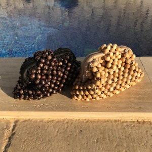 May include: Two beaded purses, one brown and one natural wood, are sitting on a light brown surface. The purses are made of small round beads that are strung together.