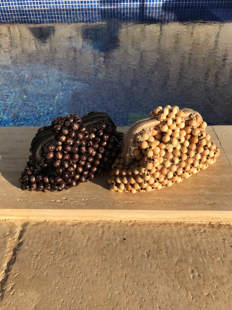 May include: Two beaded purses, one brown and one natural wood, are sitting on a light brown surface. The purses are made of small round beads that are strung together.