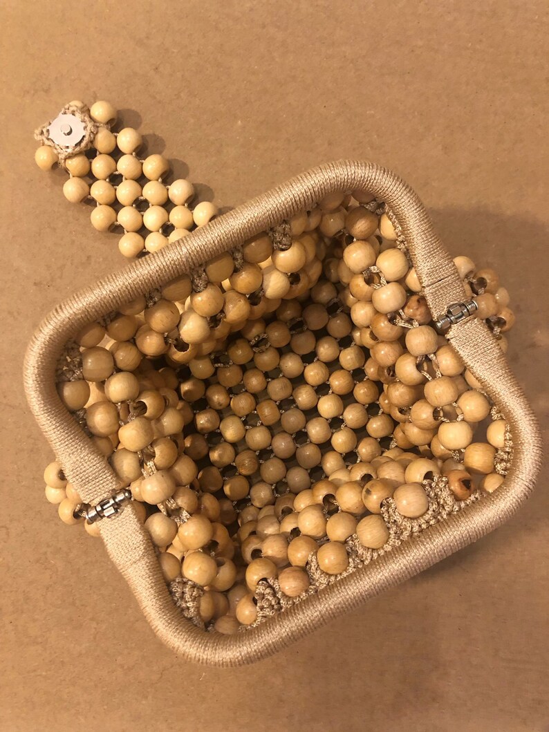 May include: A square-shaped beaded bag made of natural wood beads. The bag has a clasp closure and a beaded strap.