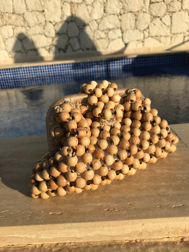 May include: A beige beaded clutch bag made of woven wood beads. The bag is sitting on a light brown surface.