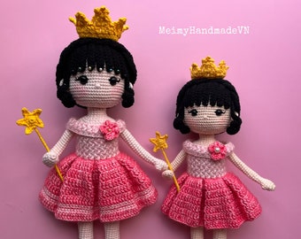 Könnte beinhalten: Zwei gehäkelte Puppen, die als Prinzessinnen verkleidet sind, tragen rosa Kleider, gelbe Kronen und halten gelbe Sternenstäbe. Die Puppen haben schwarzes Haar und rosa Wangen. Der Hintergrund ist rosa.