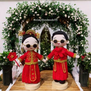 Op de afbeelding: Twee gehaakte poppen in traditionele rode trouwkleding, hand in hand. De bruidspop draagt een hoofddeksel en een bril. De bruidegompop draagt ook een bril. De poppen staan op houten voetstukken, met een bloemenboog op de achtergrond.