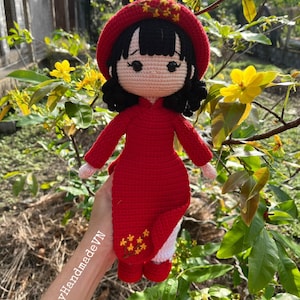 May include: A crocheted doll wearing a red dress and hat with yellow flower accents. The doll has black hair and a pink face. The doll is being held by a hand with the text "Mei my Handmade VN" written on it.