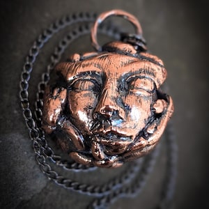 Puede incluir: Un collar con colgante de color cobre con una escultura detallada de una cara. El colgante cuelga de una cadena oscura. La cara tiene los ojos cerrados y una expresión serena. The Magickal Hud Shop está impreso en la parte inferior derecha.